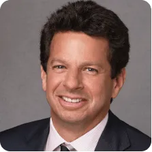 Smiling man with dark curly hair wearing a dark suit, white shirt, and striped tie against a gray background.